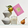 A stylized shot of Glasshouse Fragrances' A Moment in Tokyo candle and its bright pink product box sitting next to a sculpture flanked by a lemon and a pear.