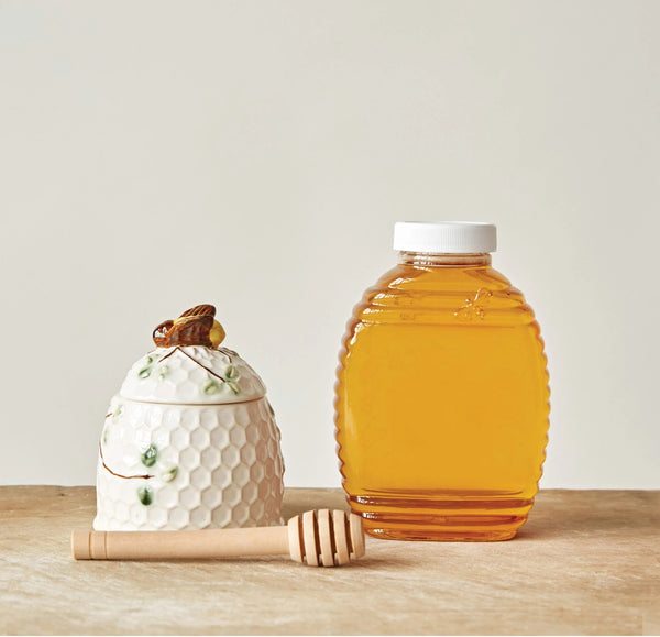 Lidded stoneware honey jar with a honeycomb texture. A hand-painted bee sits on the lid and leaves decorate the jar. Pictured with a wooden honey dipper and a jar of honey.