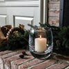 Single Simon Pearce Burlington clear rounded glass candleholder with a lit candle inside sits on a brick fireplace hearth next to a pine garland.