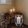 Three Simon Pearce Burlington clear rounded glass candleholders in varying sizes with lit candles inside sit atop two tables beside a sprig of cherry blossoms.