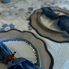 Blue linen napkin sitting on cream and blue plate, atop Navy and bone braided placemat with scalloped edge.