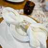 A horn napkin ring is on a cream napkin, white bowl and plate and brown patterned tablecloth with a brown glass in the background.