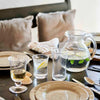 Bubble glasses and pitcher on a dark wood table set with plate, flatwares, shells, napkins with tan pillows. 