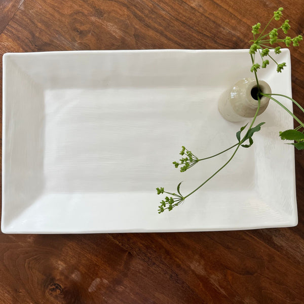 Cream, rectangle tray on wooden table with small vase and green flowers.