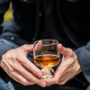 A close-up of a person holding a small bourbon glass with amber liquid in both hands, wearing a dark navy jacket.