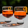 Two stemmed lowball glasses filled with an orange-red cocktail, each garnished with a curled orange peel, set on a reflective white surface with a blurred green background.