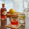 A bottle of Via Carota sits on a reflective bar counter next to two textured rocks glasses filled with cocktails, with a lemon and cutting board blurred in the background.