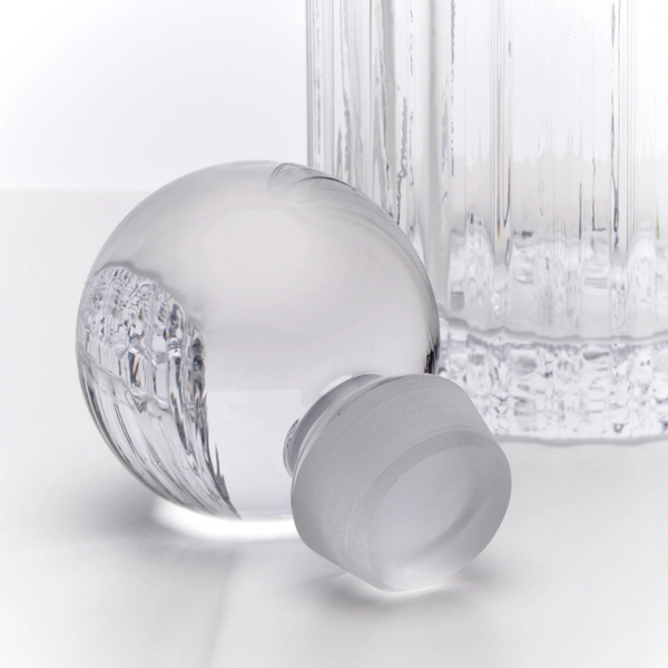 A clear glass bottle with a frosted screw cap lying on its side, with a ribbed clear glass bottle body standing behind it on a white studio background.