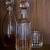 A clear ribbed glass decanter with a spherical stopper, partially filled with amber liquor, stands on a wooden surface between two matching clear glass bottles and a matching tumbler glass against a dark wood background.