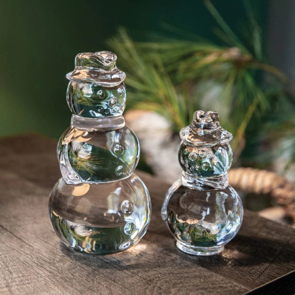 Two Simon Pearce snowmen figurines on wooden tabletop with greenery in background.