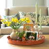 A coffee table holds an orange lacquered tray with photo frames, vases with daffodils, and two tan colored bobbin shaped candlesticks with green candles.