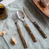 Buffalo patterned spoon, fork and knife on light blue tablecloth with bread, breadboard and glass.