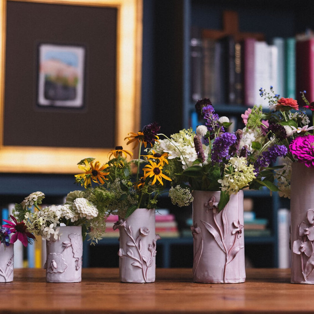 A row of five white pottery vases from Cabell's Designs, positioned from smallest to largest. Each is filled with colorful wildflowers.