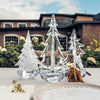 A collection of three Simon Pearce glass tabletop evergreens sit on a table with the Simon Pearce factory in the background.