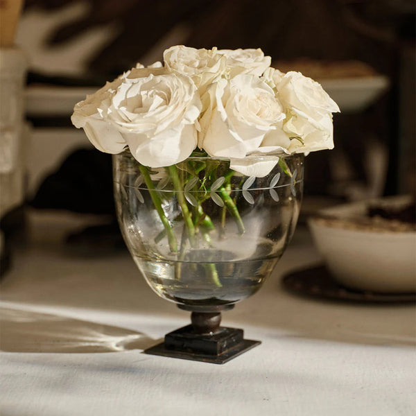 Clear glass vase on iron stand filled with white roses on a table.