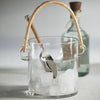 Clear glass ice bucket with rattan handle holding ice and silver ice scoop.