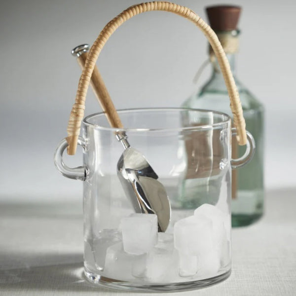 Clear glass ice bucket with rattan handle holding ice and silver ice scoop.