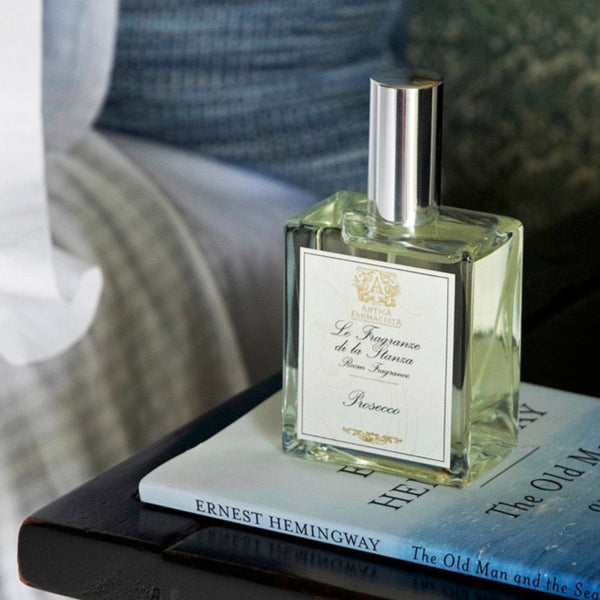 Bottle of Prosecco scented room spray on a book sitting on a nightstand next to a bed.