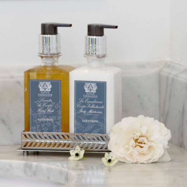 Bottle of Santorini soap and lotion in a silver tray on a marble countertop with flowersand mirror.