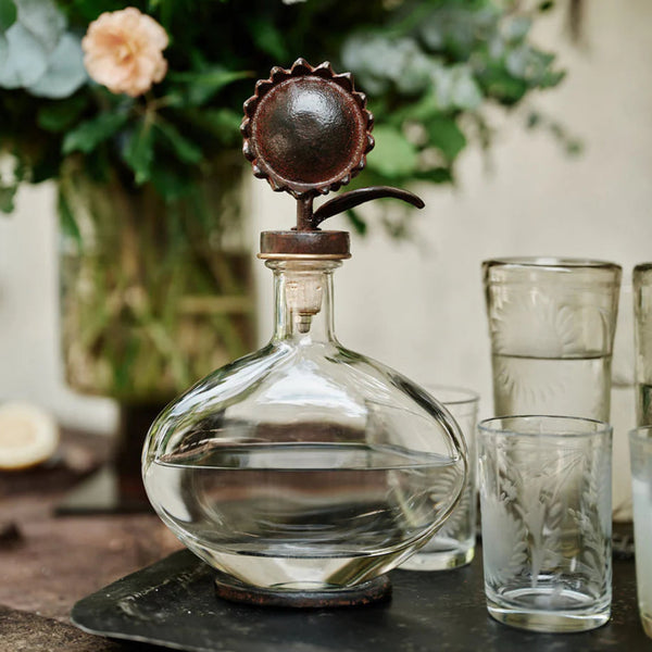 Clear glass decanter with sunflower stopper on a table with glasses and flowers in the background
