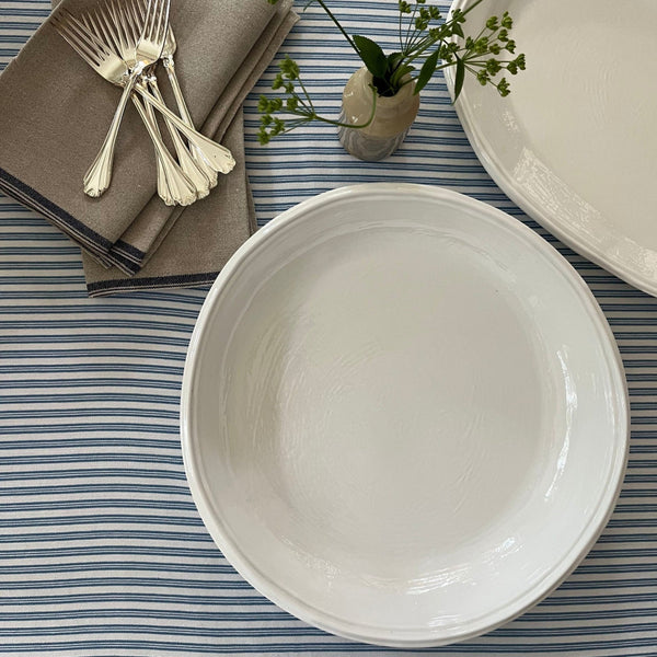 Double Line Cream bowl and platter on striped table cloth with linen napkin, forks and vase with green flowers. 