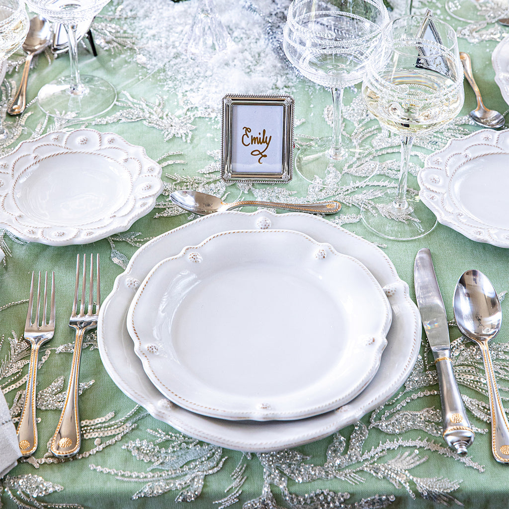 White Juliska Berry & Thread flared plates, set on a table with a green tablecloth, glassware and silverware. A small silver frame holds a card for the guest's name. 