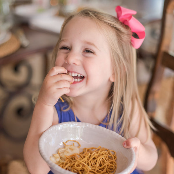 A smiling girl with a pink bow in her hair eats spaghetti from a Relish melamine bowl with a beaded edge.