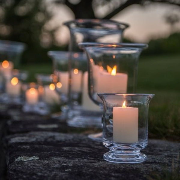 Various clear glass Simon Pearce candleholders lined up on a wall outside. It is evening and there are lit candles placed inside.