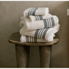 Two white Mungo waffle weave bath towels and two hand towels folded and stacked on a wooden stool. The towels have two thick and two thin gray stripes near the fringed edge.