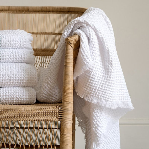 Four Mungo white waffle weave bath towels folded and stacked on a wicker bench. One white waffle towel is draped over the back of the bench.