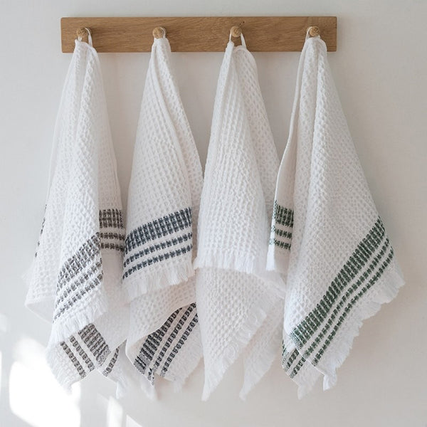 Four Mungo waffle weave hand towels hanging on pegs. One towel has gray stripes near the fringed edge, one has blue stripes, one is solid white, and one has green stripes.