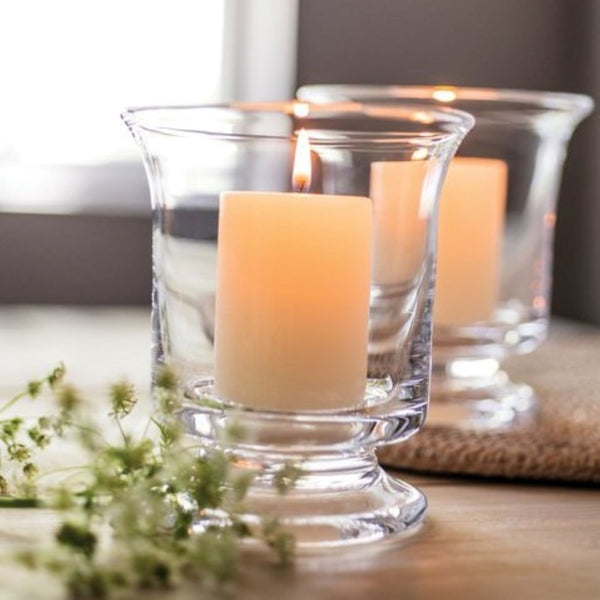 Pair of small Simon Pearce Revere Hurricanes on a table with lit candles placed inside. These clear glass candleholders are 5.25 inches tall and 4.5 inches wide and have a sturdy flared base and flared top.
