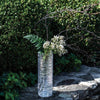Tall Simon Pearce Echo Lake vase filled with branches, ferns and flowers sits atop a stone wall.