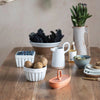 White fluted stoneware bowls, square and round, hold kiwi fruit and blueberries and sit on a counter with other white kitchen serving pieces.