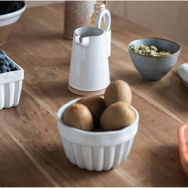 A white fluted stoneware bowl filled with kiwi fruit sits on a counter with other white kitchen serving pieces.