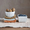 A round white fluted stoneware bowl filled with kiwi fruit and a square fluted stoneware bowl filled with blueberries sit atop napkins and a tablecloth.