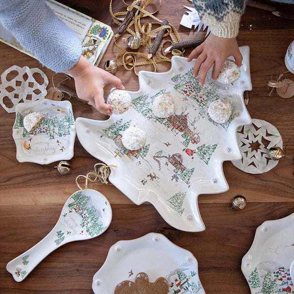 Juliska's tree-shaped platter from the North Pole collection sits on a holiday table and holds several cookies. Children's hands are seen reaching for some of the cookies. 