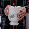 Juliska's North Pole mug is decorated with a wintery scene and filled with hot cocoa and a candy cane garnish. It's held by a person in a plaid shirt.