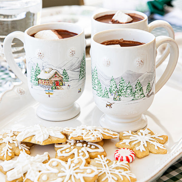 Two Juliska North Pole mugs decorated with a wintery scene are filled with hot cocoa and marshmallows and sit on a table alongside a plate of cookies.