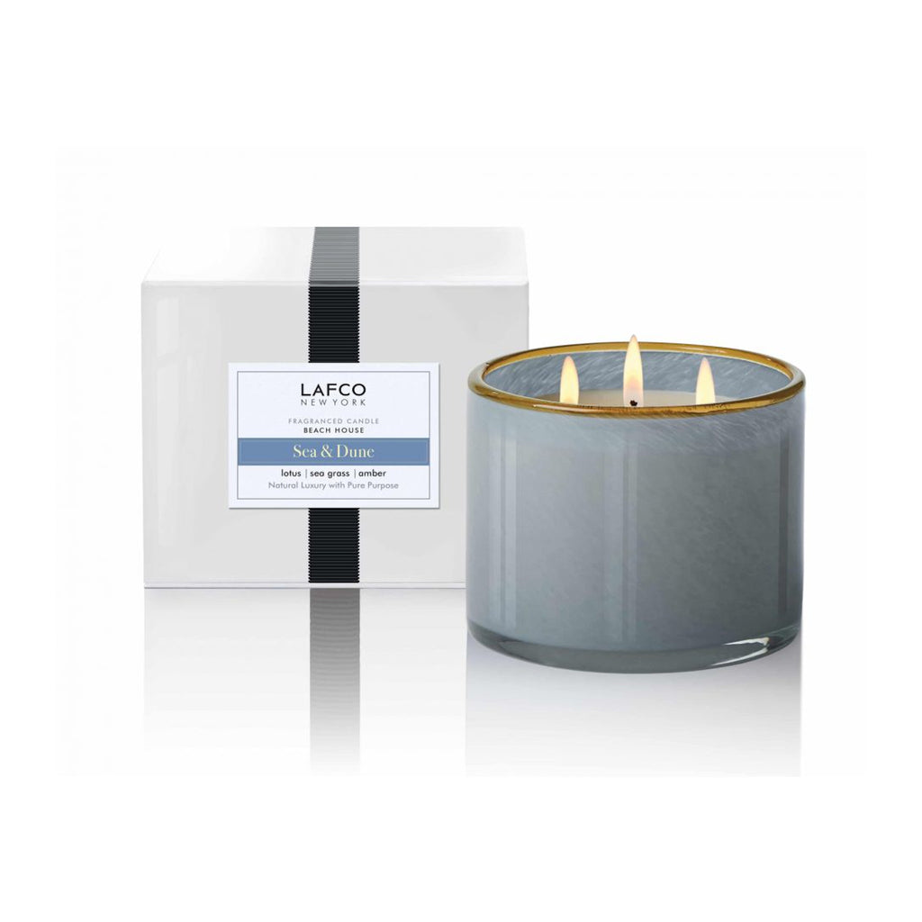 Lafco's Sea & Dune candle sits alongside its signature gift box. The candle is housed in a hand-blown blue glass vessel with a honey-colored rim. The candle's three wicks are lit.