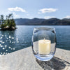 Single Simon Pearce Burlington clear rounded glass candleholder with a candle inside sits on a stone ledge overlooking a lake on a sunny day.